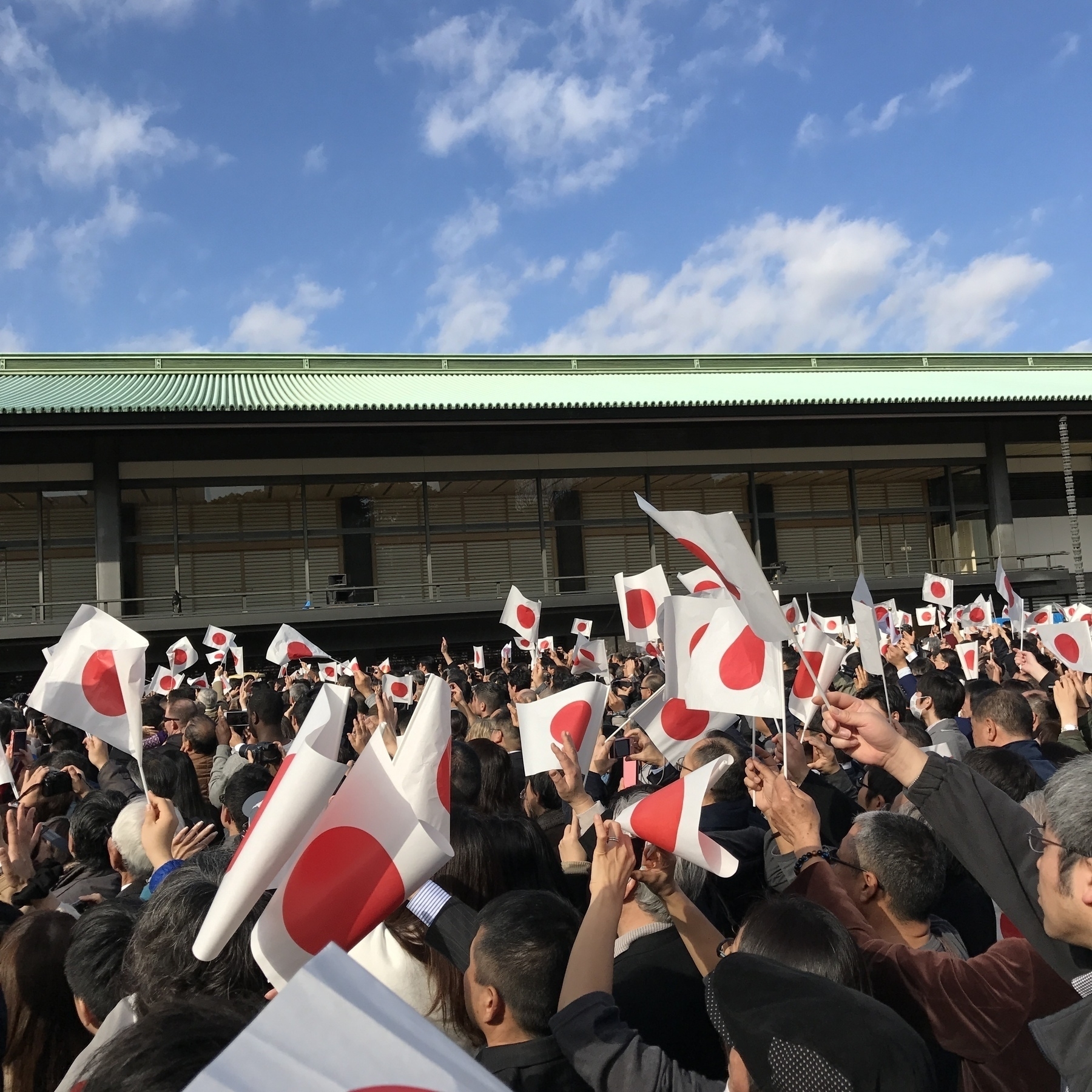 A large crowd of people wave paper Japanese flags outside of Tokyo Imperial Palace.