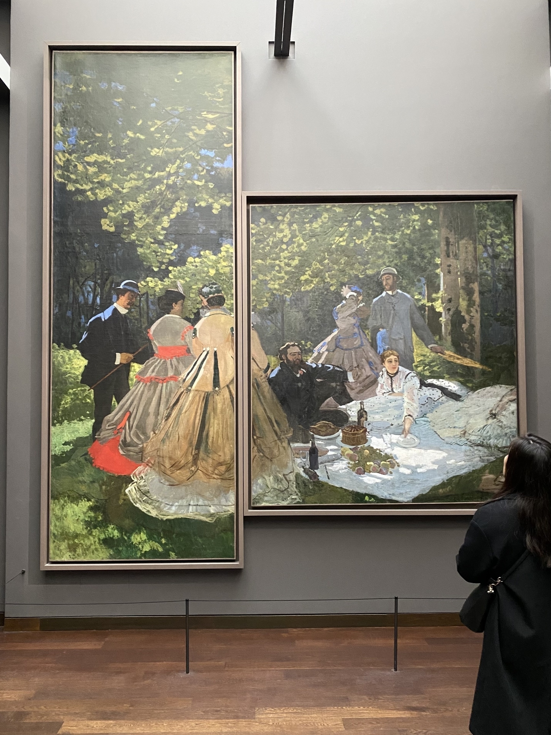 A museum visitor looks up at a two segment painting. The left frame is a tall rectangle. The right frame, connecting midway down, is a more typical portrait sized canvas. Together these impressionist oil paintings capture a picnic under a beautiful tree.